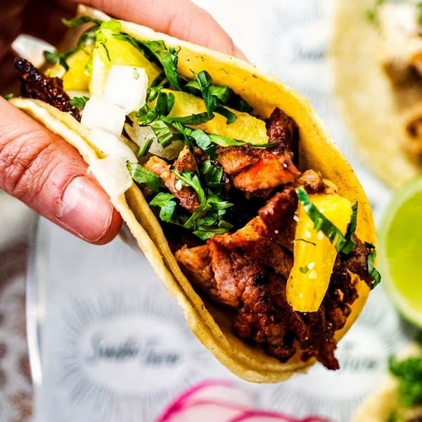 Hand holding a taco filled with meat, pineapple, cilantro, and onions, with a lime in the background.