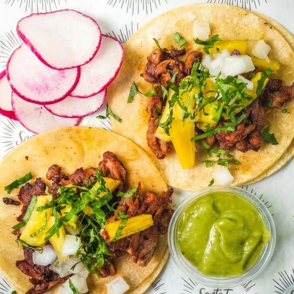 Two Santo Taco al pastor tacos with pineapple, onions, cilantro, radishes, and guacamole.