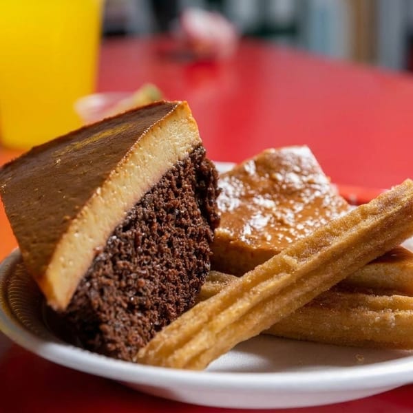 A plate of chocolate flan cake and churros on a red table with a drink in the background.