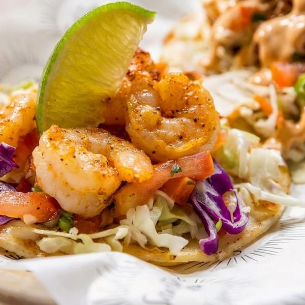 Two shrimp tacos with cabbage, pico de gallo, and a lime wedge on branded paper.
