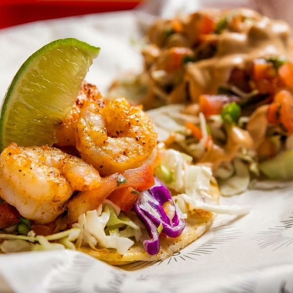 Two shrimp tacos with cabbage, tomatoes, and lime wedges on branded paper.