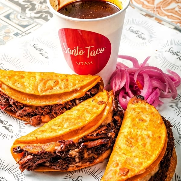 Three Santo Taco birria tacos with a cup of consommé and pickled red onions on branded paper.