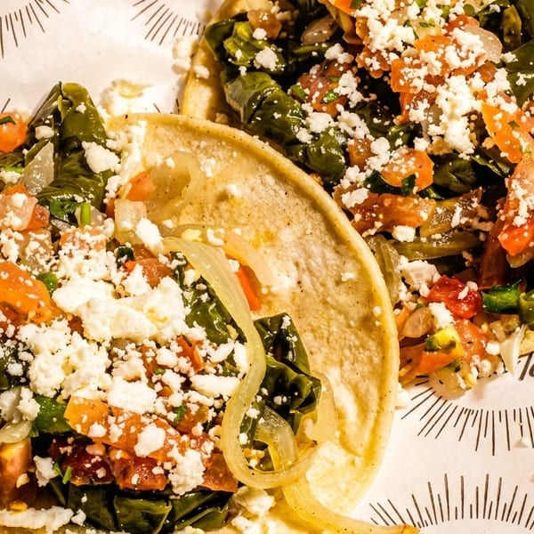 Two vibrant tacos filled with green vegetables, tomatoes, onions, and crumbled white cheese on patterned paper.