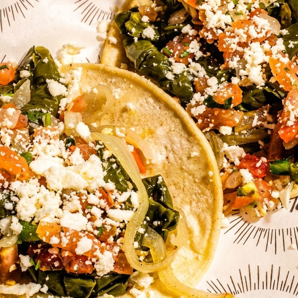 Two vibrant tacos filled with green vegetables, tomatoes, onions, and crumbled white cheese on patterned paper.